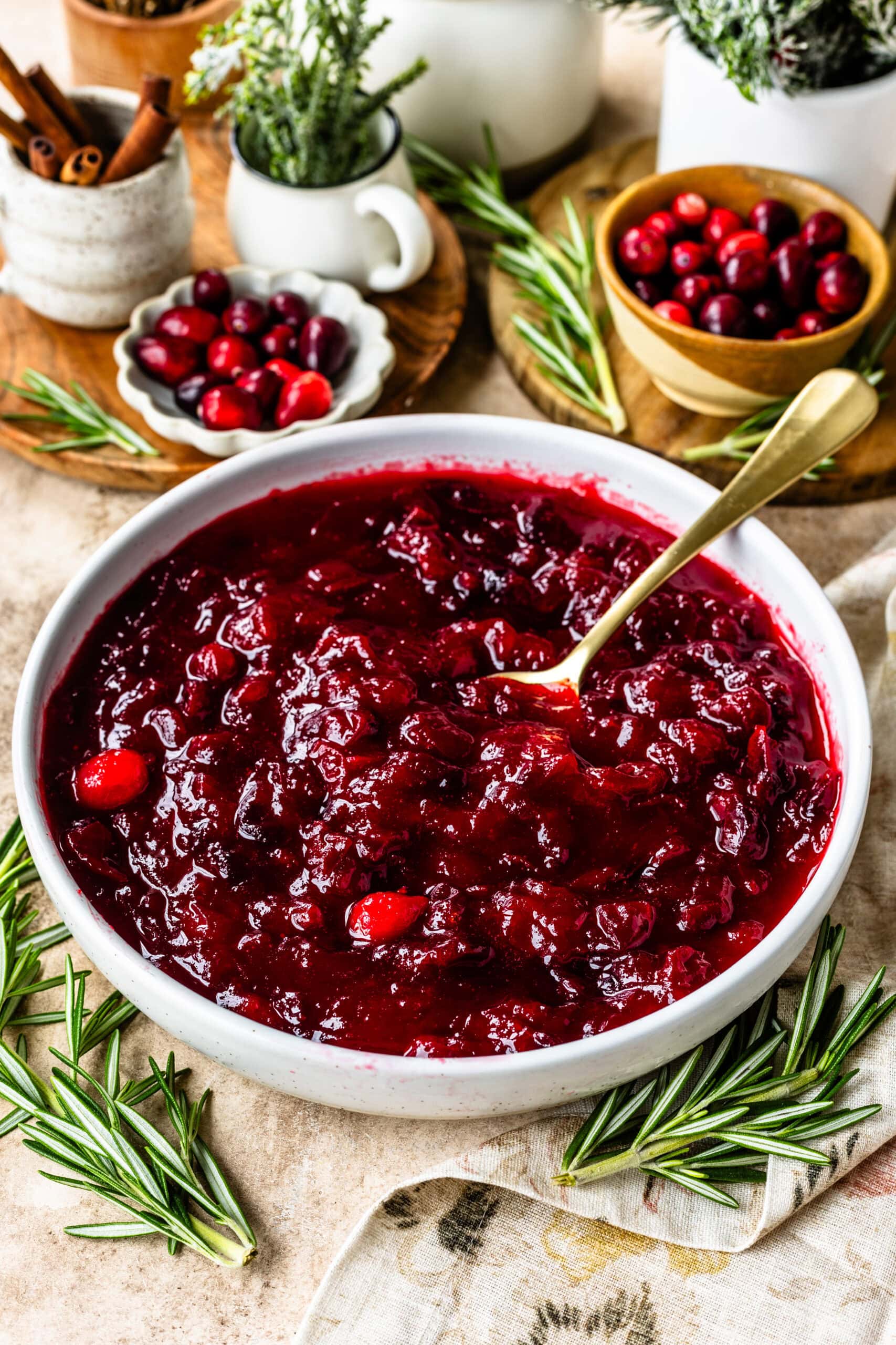 The image shows a bowl of rich, vibrant cranberry sauce with a deep red color, indicating a thick and flavorful texture. A gold spoon rests in the sauce, ready for serving. Surrounding the bowl are fresh rosemary sprigs, adding a touch of greenery and a festive feel to the scene. In the background, small bowls hold extra cranberries and cinnamon sticks, contributing to a cozy, holiday ambiance. The setup includes rustic wooden elements and greenery, creating a warm and inviting presentation perfect for a seasonal spread.