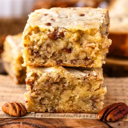 Close-up of two stacked pecan blondie squares on a wooden board with music sheet details, highlighting their golden, moist texture and visible pecan pieces, surrounded by pecans and white chocolate chunks.