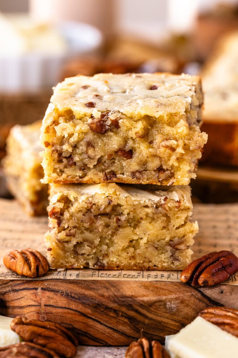 Close-up of two stacked pecan blondie squares on a wooden board with music sheet details, highlighting their golden, moist texture and visible pecan pieces, surrounded by pecans and white chocolate chunks.