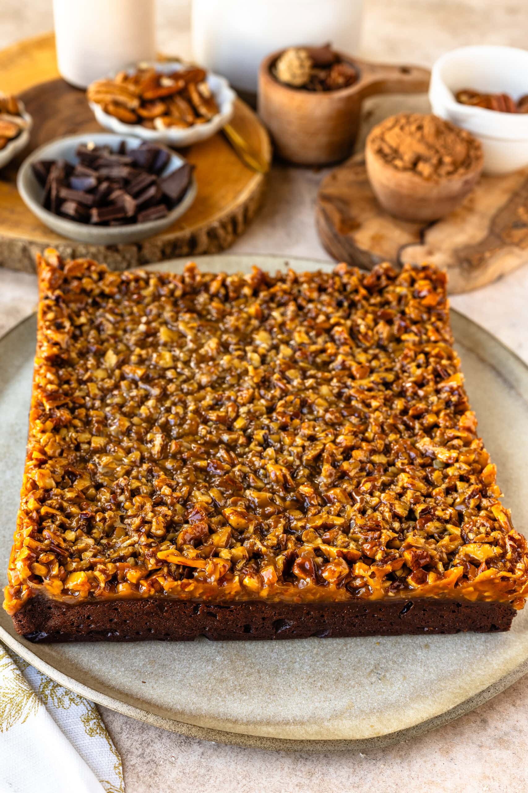 A full square of pecan pie brownies on a rustic ceramic plate, showcasing the perfectly even layers. The top layer is a golden, caramelized pecan topping, rich and textured, spread smoothly across the fudgy brownie base. In the background, ingredients like chopped chocolate, pecans, and cocoa powder are arranged on wooden boards, adding a cozy, homemade feel to the scene. This dessert is a delicious blend of flavors and textures, perfect for any special occasion or gathering.