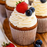 A close up of a golden cupcake topped with creamy stabilized whipped cream swirls, garnished with plump blueberries and fresh strawberries, sitting on a wooden board for a delightful presentation.