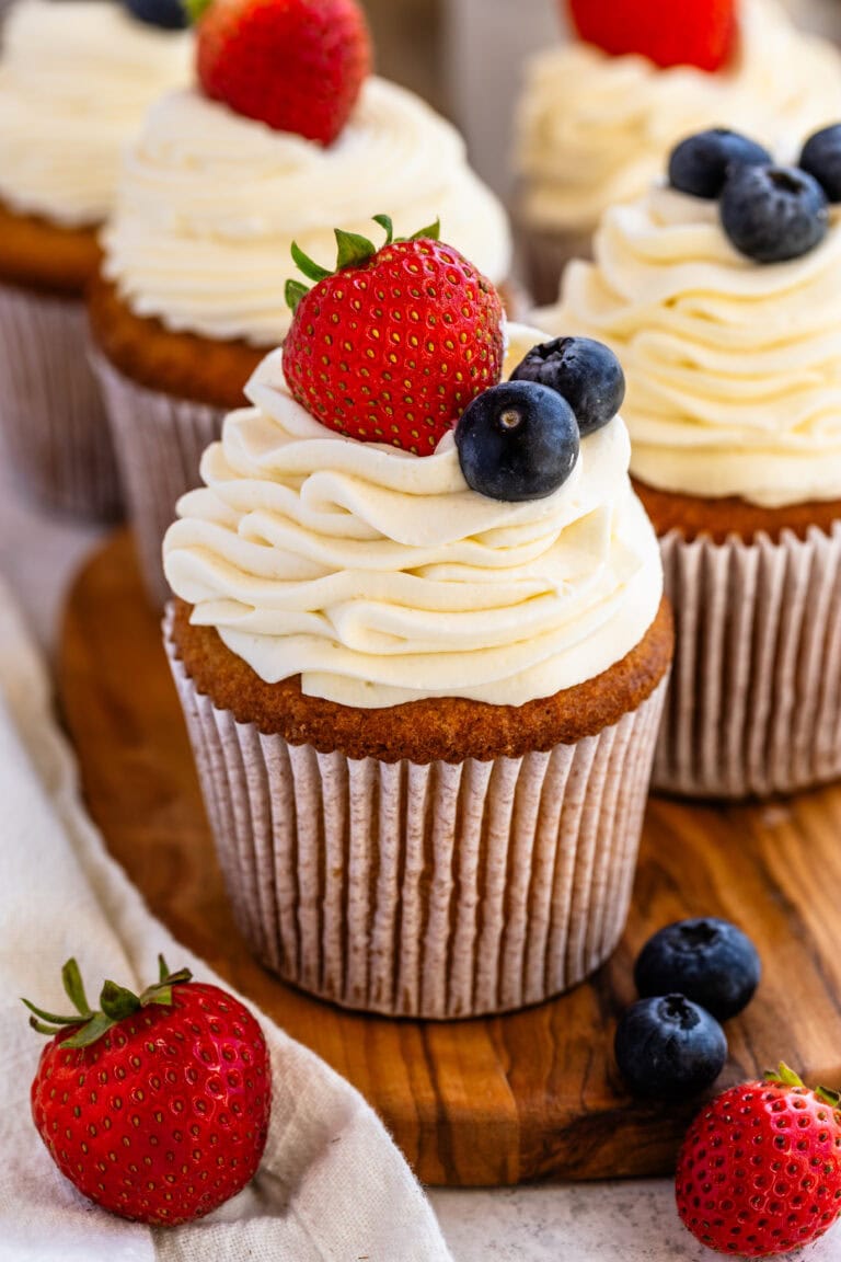 A close up of a golden cupcake topped with creamy stabilized whipped cream swirls, garnished with plump blueberries and fresh strawberries, sitting on a wooden board for a delightful presentation.