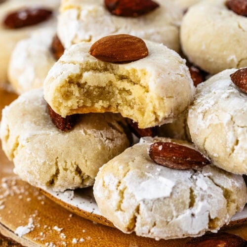 close-up of almond cookies dusted with powdered sugar, each topped with a whole almond, with one cookie showing a bite taken out to reveal a soft and crumbly interior.