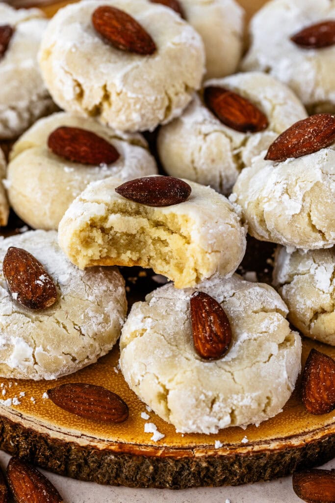 close-up of almond cookies dusted with powdered sugar, each topped with a whole almond, with one cookie showing a bite taken out to reveal a soft and crumbly interior.