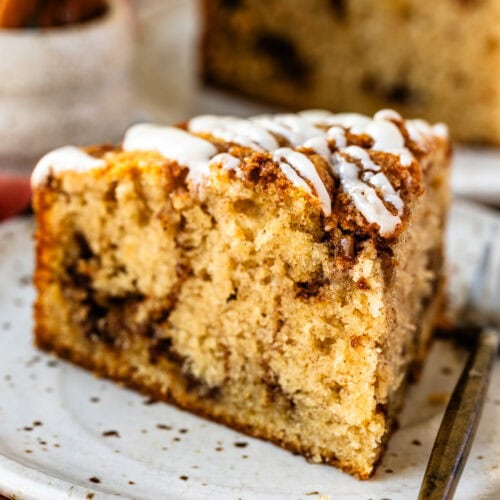 A close up of a slice of cinnamon roll cake sits on a speckled ceramic plate, showcasing its moist crumb, cinnamon swirls, and drizzled icing on top. The remaining cake is visible in the background on a wooden board, surrounded by warm, cozy cinnamon sticks and rustic decor.