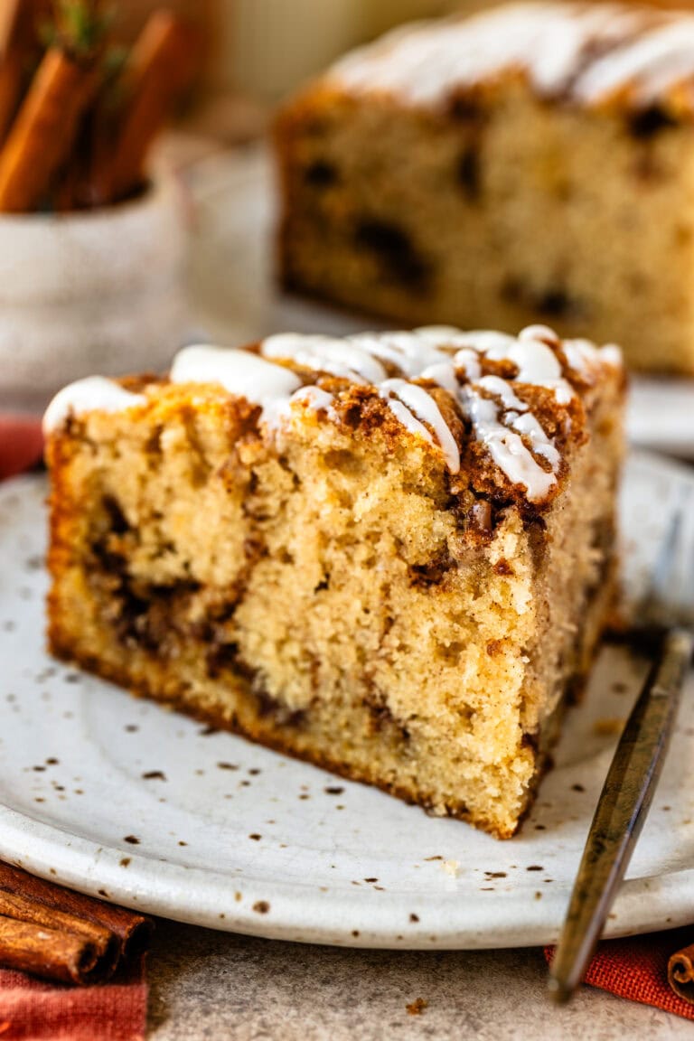 A close up of a slice of cinnamon roll cake sits on a speckled ceramic plate, showcasing its moist crumb, cinnamon swirls, and drizzled icing on top. The remaining cake is visible in the background on a wooden board, surrounded by warm, cozy cinnamon sticks and rustic decor.
