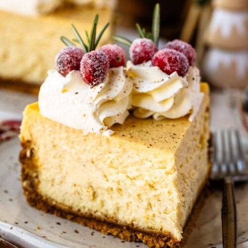 A close-up slice of creamy eggnog cheesecake on a plate, topped with whipped cream, sugared cranberries, and a sprig of rosemary for festive garnish.