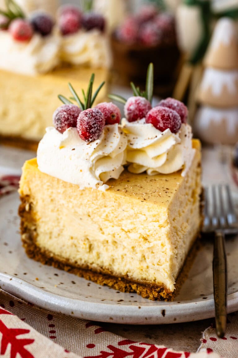 A close-up slice of creamy eggnog cheesecake on a plate, topped with whipped cream, sugared cranberries, and a sprig of rosemary for festive garnish.