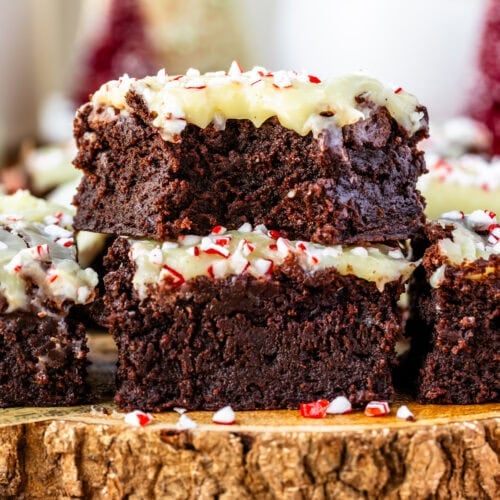 A stack of peppermint brownies topped with white chocolate ganache and crushed candy canes. The brownies have a rich, fudgy texture, with vibrant red and white peppermint pieces adding a festive holiday touch. Candy canes are scattered around the rustic wooden serving board for a cozy, seasonal vibe.