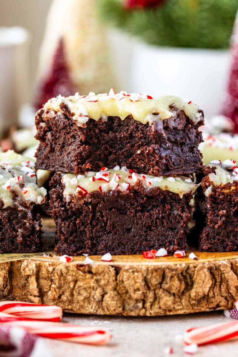A stack of peppermint brownies topped with white chocolate ganache and crushed candy canes. The brownies have a rich, fudgy texture, with vibrant red and white peppermint pieces adding a festive holiday touch. Candy canes are scattered around the rustic wooden serving board for a cozy, seasonal vibe.