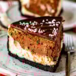 A slice of peppermint cheesecake on a plate. The dessert features a rich chocolate cookie crust, layers of peppermint-flavored cheesecake and chocolate filling, topped with glossy chocolate ganache and sprinkled with crushed candy canes. Candy canes and festive decorations are visible in the background.