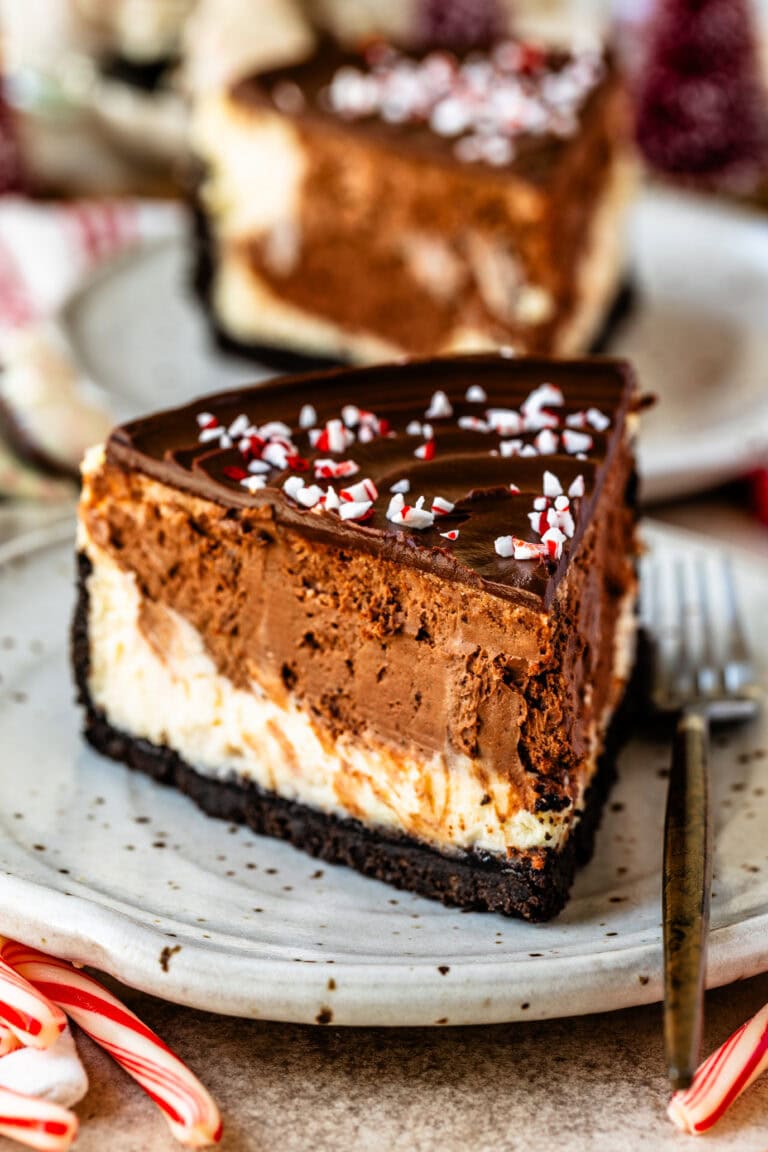 A slice of peppermint cheesecake on a plate. The dessert features a rich chocolate cookie crust, layers of peppermint-flavored cheesecake and chocolate filling, topped with glossy chocolate ganache and sprinkled with crushed candy canes. Candy canes and festive decorations are visible in the background.