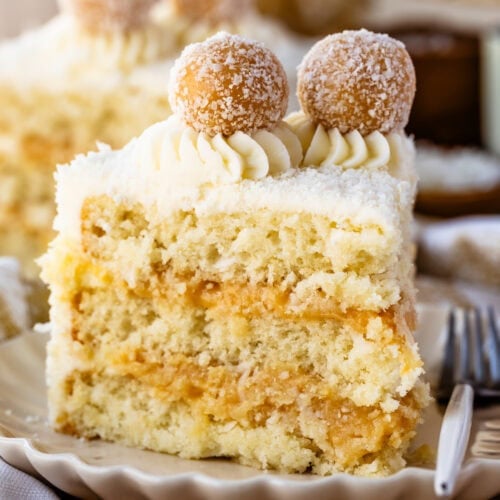 A slice of coconut cake with layers of fluffy coconut sponge and creamy filling, topped with piped frosting and two coconut-covered truffles. The cake sits on a scalloped plate, creating a cozy and festive presentation.