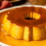 A close-up of a Spanish flan with a rich caramel glaze, set on a white plate with a decorative fluted edge. The golden, creamy texture of the flan contrasts beautifully with the glossy caramel sauce pooling around it, creating an elegant presentation.
