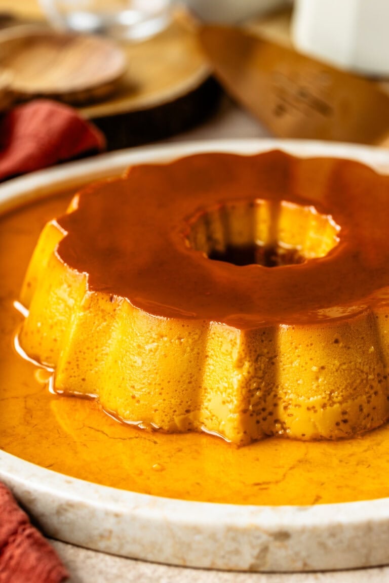 A close-up of a Spanish flan with a rich caramel glaze, set on a white plate with a decorative fluted edge. The golden, creamy texture of the flan contrasts beautifully with the glossy caramel sauce pooling around it, creating an elegant presentation.