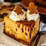 Close-up of a slice of gingerbread cheesecake on a decorative white plate. The cheesecake is topped with whipped cream, gingerbread cookie decorations shaped like a star and a gingerbread man, and drizzled with caramel sauce. The background includes blurred holiday-themed elements and gingerbread cookies, enhancing the festive presentation.