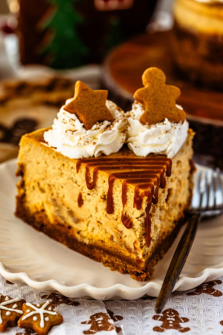 Close-up of a slice of gingerbread cheesecake on a decorative white plate. The cheesecake is topped with whipped cream, gingerbread cookie decorations shaped like a star and a gingerbread man, and drizzled with caramel sauce. The background includes blurred holiday-themed elements and gingerbread cookies, enhancing the festive presentation.