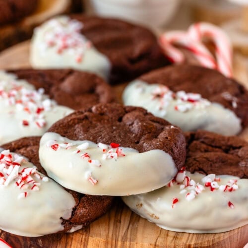 Chocolate peppermint cookies dipped in white chocolate and topped with crushed candy canes, stacked on a wooden board with festive holiday decorations in the background.