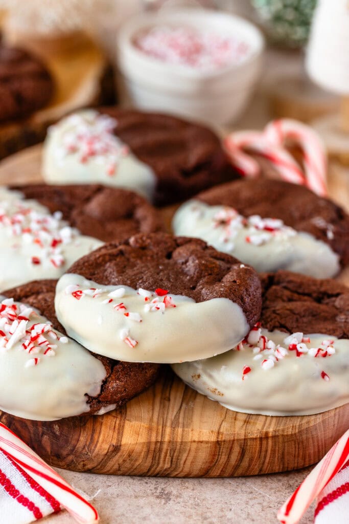 Chocolate peppermint cookies dipped in white chocolate and topped with crushed candy canes, stacked on a wooden board with festive holiday decorations in the background.