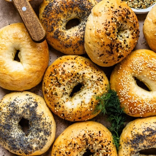 Golden brown bagels topped with a variety of seeds and seasonings, including poppy seeds, sesame seeds, and everything bagel seasoning, arranged on a rustic surface with a bowl of cream cheese and a knife in the background.
