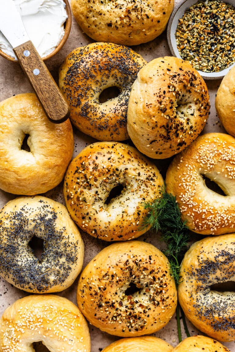 Golden brown bagels topped with a variety of seeds and seasonings, including poppy seeds, sesame seeds, and everything bagel seasoning, arranged on a rustic surface with a bowl of cream cheese and a knife in the background.