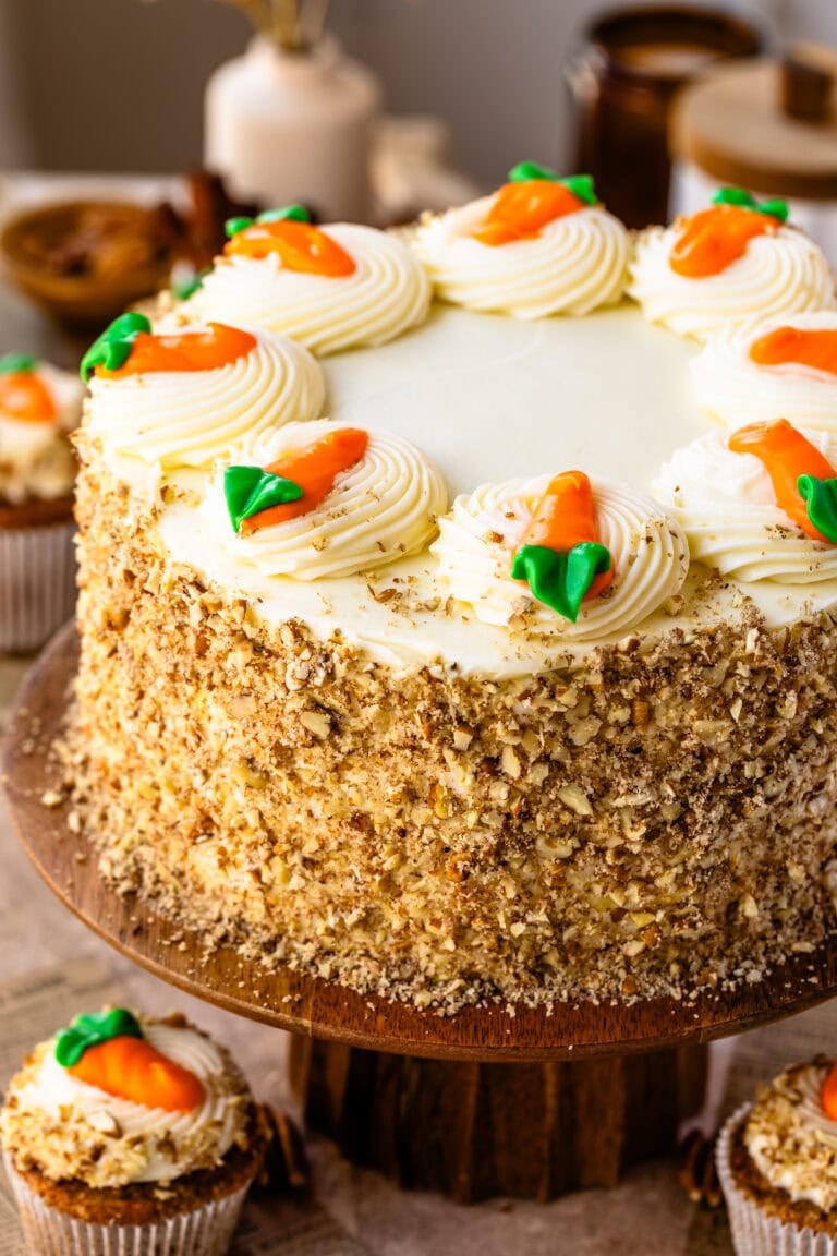 A whole carrot cake displayed on a wooden cake stand, frosted with cream cheese frosting and decorated with piped swirls topped with frosting carrots. The sides of the cake are coated with chopped nuts, and a single carrot-topped cupcake is visible at the base, with a blurred background featuring jars and décor.