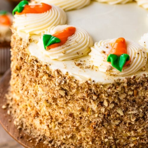 A close up of a whole carrot cake displayed on a wooden cake stand, frosted with cream cheese frosting and decorated with piped swirls topped with frosting carrots. The sides of the cake are coated with chopped nuts, and a single carrot-topped cupcake is visible at the base, with a blurred background featuring jars and dรฉcor.