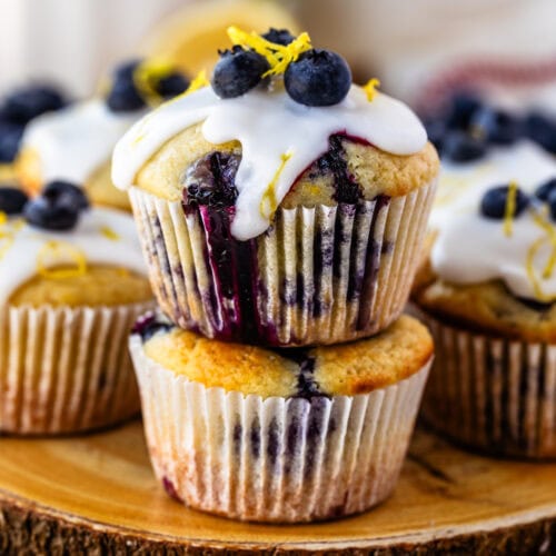 Lemon blueberry muffins topped with a drizzle of icing, fresh blueberries, and lemon zest, served on a rustic wooden platter.