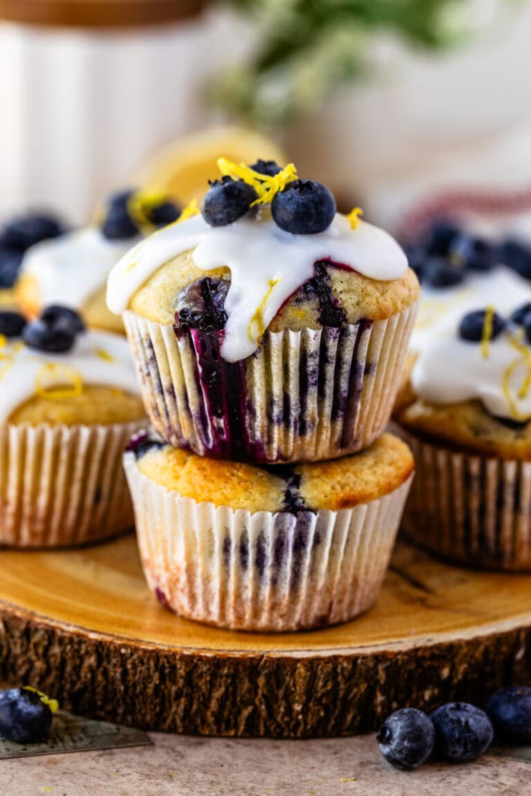 Lemon blueberry muffins topped with a drizzle of icing, fresh blueberries, and lemon zest, served on a rustic wooden platter.