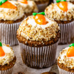 A close-up view of beautifully decorated carrot cupcakes, topped with cream cheese frosting, crushed nuts, and an adorable candy carrot garnish. The cupcakes are arranged on a rustic surface with pecans scattered around.