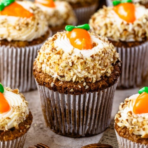 A close-up view of beautifully decorated carrot cupcakes, topped with cream cheese frosting, crushed nuts, and an adorable candy carrot garnish. The cupcakes are arranged on a rustic surface with pecans scattered around.