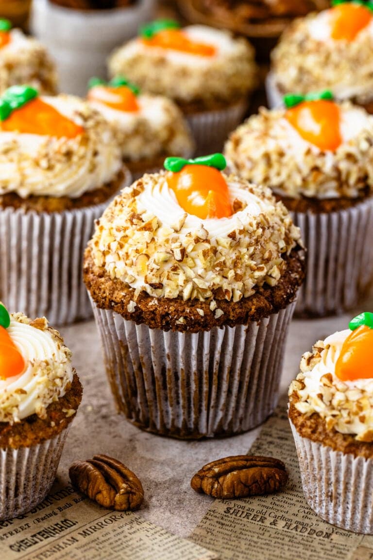A close-up view of beautifully decorated carrot cupcakes, topped with cream cheese frosting, crushed nuts, and an adorable candy carrot garnish. The cupcakes are arranged on a rustic surface with pecans scattered around.
