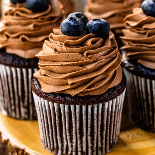 A chocolate cupcake topped with swirls of chocolate whipped cream frosting and fresh blueberries. Other frosted cupcakes with blueberries are visible in the background.