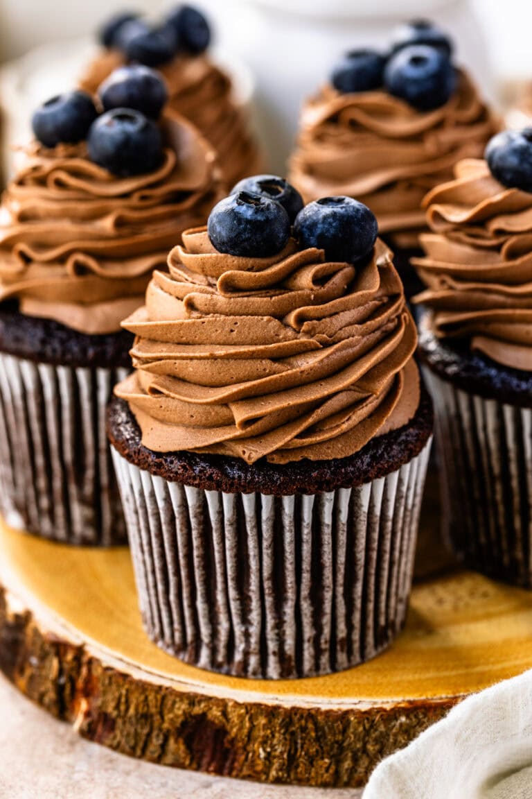 A chocolate cupcake topped with swirls of chocolate whipped cream frosting and fresh blueberries. Other frosted cupcakes with blueberries are visible in the background.