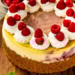 Raspberry cheesecake on a wooden board topped with piped whipped cream swirls and fresh raspberries, showcasing a golden crust and swirls of raspberry filling.