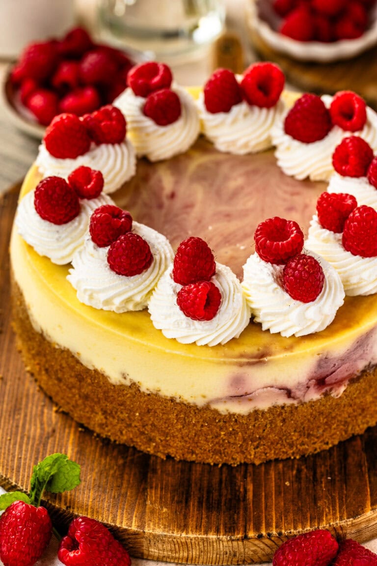 Raspberry cheesecake on a wooden board topped with piped whipped cream swirls and fresh raspberries, showcasing a golden crust and swirls of raspberry filling.