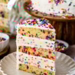 A slice of Funfetti Ice Cream Cake on a plate, showcasing layers of colorful sprinkle-filled cake and creamy vanilla ice cream, topped with whipped cream and extra sprinkles.