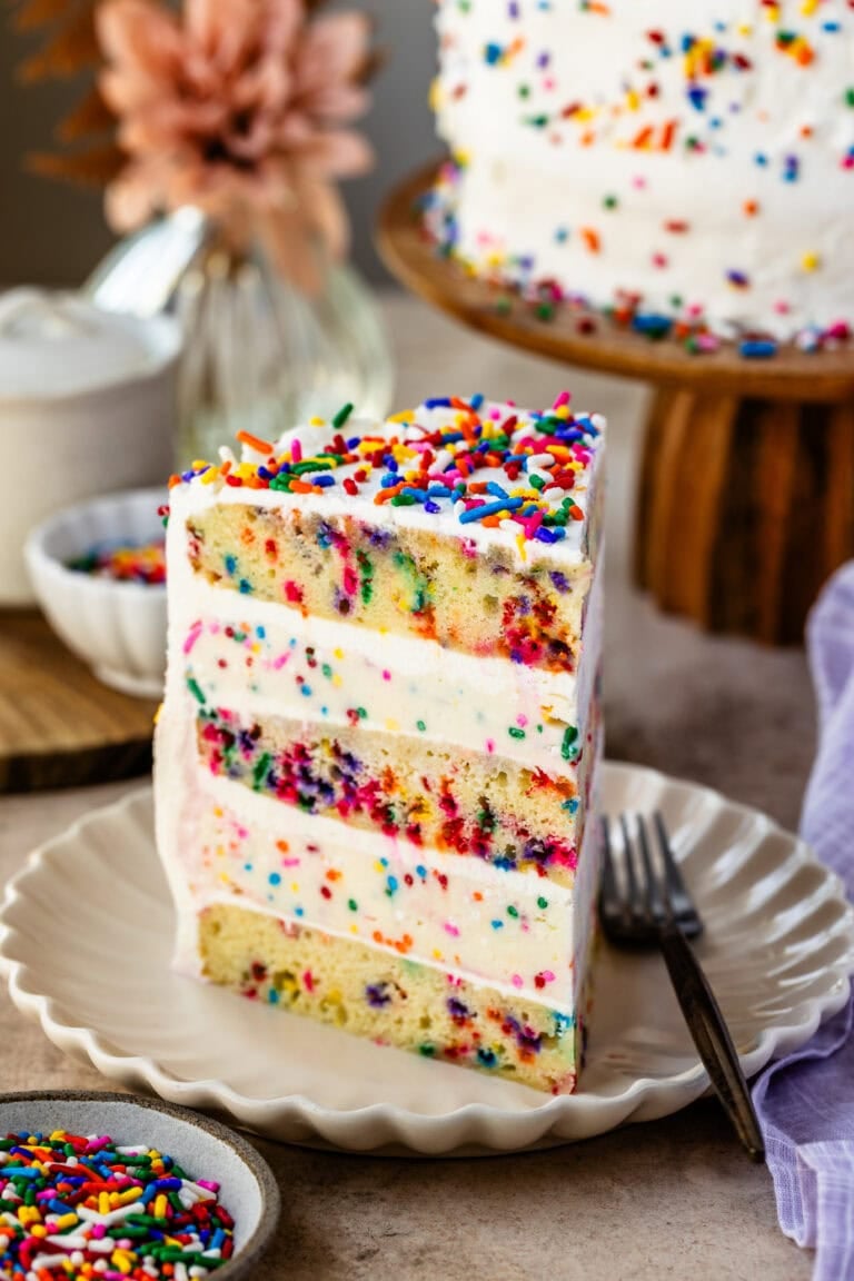 A slice of Funfetti Ice Cream Cake on a plate, showcasing layers of colorful sprinkle-filled cake and creamy vanilla ice cream, topped with whipped cream and extra sprinkles.