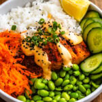 A close up of a vibrant salmon rice bowl served in a white dish on a wooden cutting board. The bowl contains air-fried salmon topped with creamy sriracha mayo, black sesame seeds, and sliced green onions, alongside fluffy white rice, shredded carrots, fresh cucumber slices, and bright green edamame. A lemon wedge sits on the side for an extra pop of freshness.