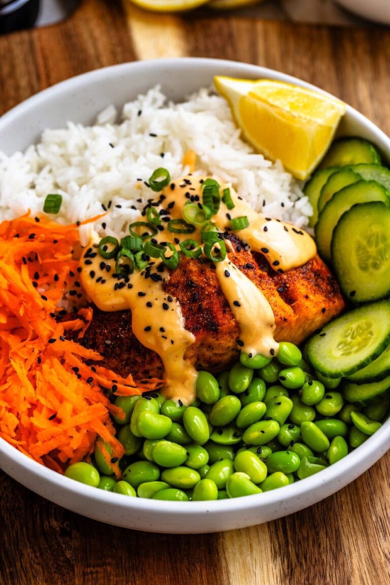 A close up of a vibrant salmon rice bowl served in a white dish on a wooden cutting board. The bowl contains air-fried salmon topped with creamy sriracha mayo, black sesame seeds, and sliced green onions, alongside fluffy white rice, shredded carrots, fresh cucumber slices, and bright green edamame. A lemon wedge sits on the side for an extra pop of freshness.