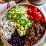 A vibrant beef bowl served in a rustic ceramic bowl, featuring seasoned ground beef, fluffy cilantro rice, black beans, shredded lettuce, cherry tomatoes, jalapeño slices, and diced avocado, all drizzled with a creamy green sauce. A lime wedge and fresh cilantro garnish the scene, with additional lime slices and sauce in the background.