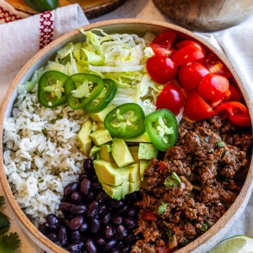 A vibrant beef bowl served in a rustic ceramic bowl, featuring seasoned ground beef, fluffy cilantro rice, black beans, shredded lettuce, cherry tomatoes, jalapeño slices, and diced avocado, all drizzled with a creamy green sauce. A lime wedge and fresh cilantro garnish the scene, with additional lime slices and sauce in the background.