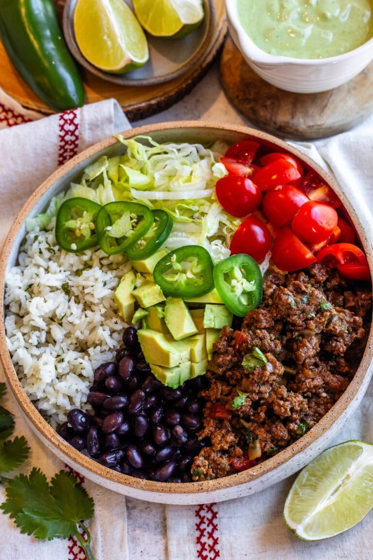 A vibrant beef bowl served in a rustic ceramic bowl, featuring seasoned ground beef, fluffy cilantro rice, black beans, shredded lettuce, cherry tomatoes, jalapeño slices, and diced avocado, all drizzled with a creamy green sauce. A lime wedge and fresh cilantro garnish the scene, with additional lime slices and sauce in the background.