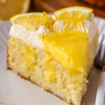 A close-up of a slice of lemon curd cake on a white plate, featuring a moist lemon-infused cake base topped with glossy lemon curd, piped whipped cream, and garnished with fresh lemon slices and zest. A fork rests beside the slice, with blurred lemons and the remaining cake in the background.