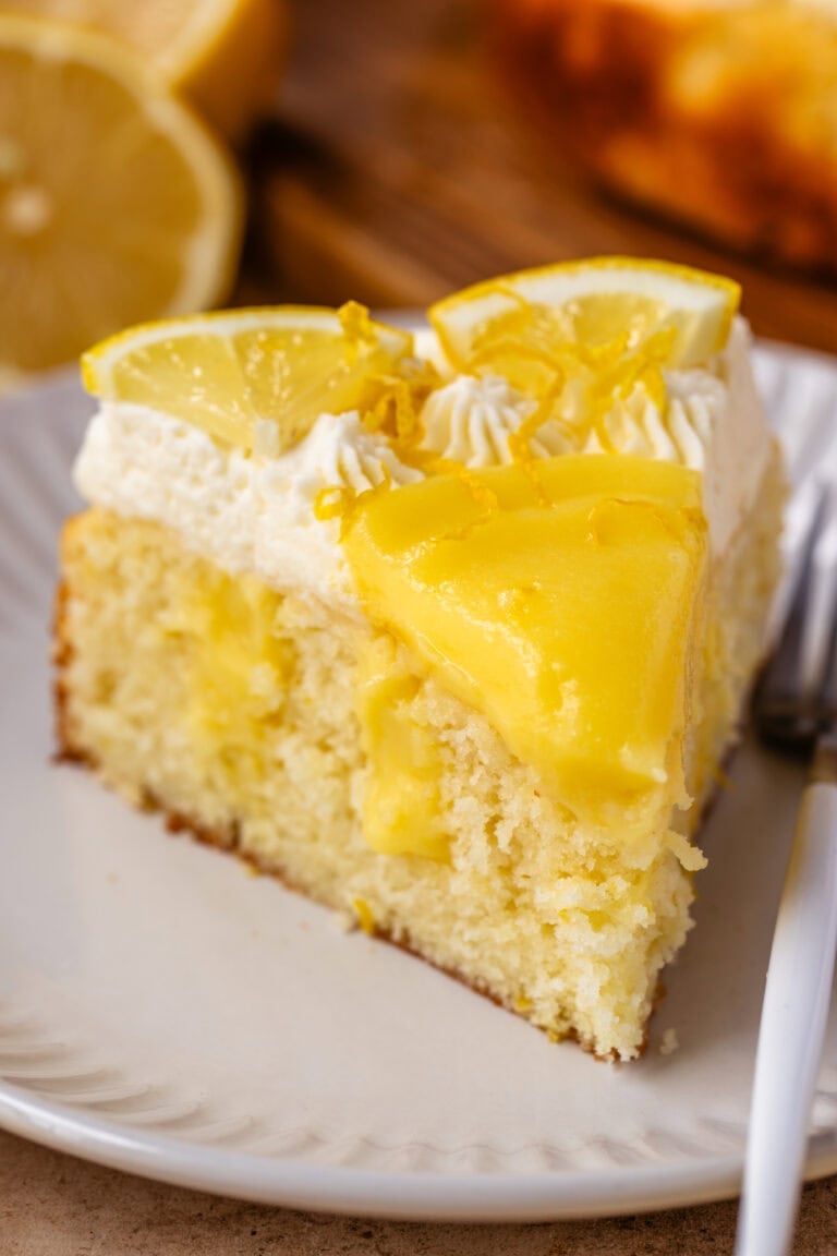 A close-up of a slice of lemon curd cake on a white plate, featuring a moist lemon-infused cake base topped with glossy lemon curd, piped whipped cream, and garnished with fresh lemon slices and zest. A fork rests beside the slice, with blurred lemons and the remaining cake in the background.
