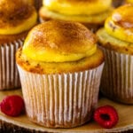 A close-up of a crème brûlée cupcake with a golden caramelized sugar topping, sitting on a wooden board with fresh raspberries nearby. More cupcakes with glossy, torched sugar tops are blurred in the background.