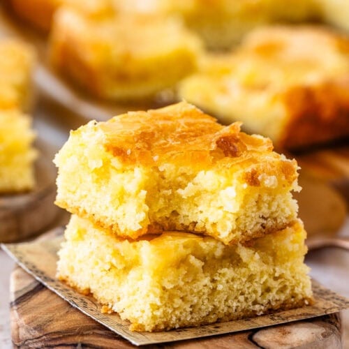 A close-up shot of two stacked pieces of gooey butter cake, showing the golden, slightly crisp top and the soft, moist interior. The cake pieces rest on a rustic wooden board with more cake pieces blurred in the background.