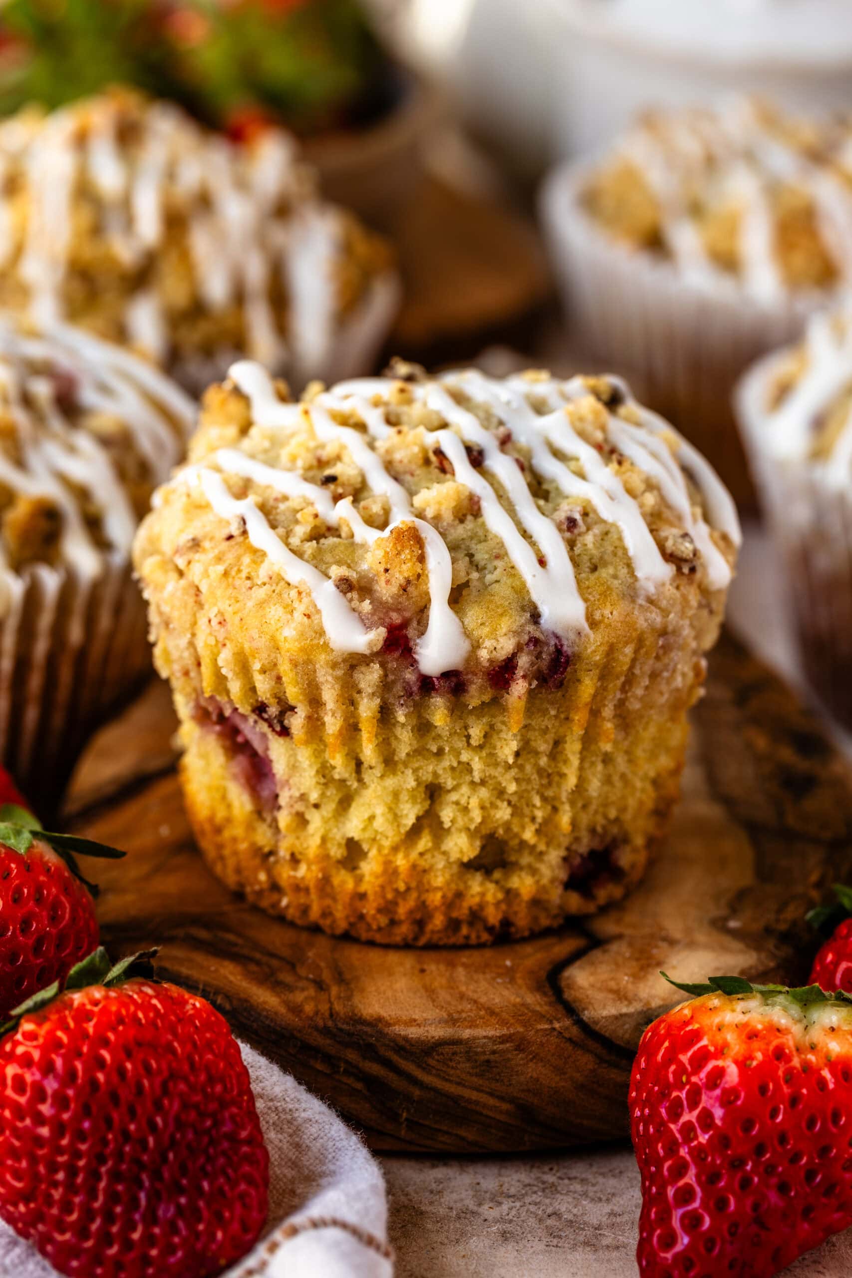 A close-up of a golden strawberry muffin with a crumbly streusel topping and a drizzle of icing. Fresh strawberries are scattered around, adding a pop of red, with more muffins blurred in the background.