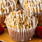 A close-up of a golden strawberry muffin with a crumbly streusel topping and a drizzle of icing. Fresh strawberries are scattered around, adding a pop of red, with more muffins blurred in the background.