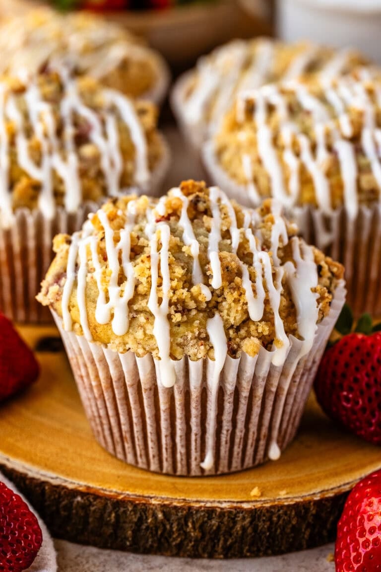A close-up of a golden strawberry muffin with a crumbly streusel topping and a drizzle of icing. Fresh strawberries are scattered around, adding a pop of red, with more muffins blurred in the background.